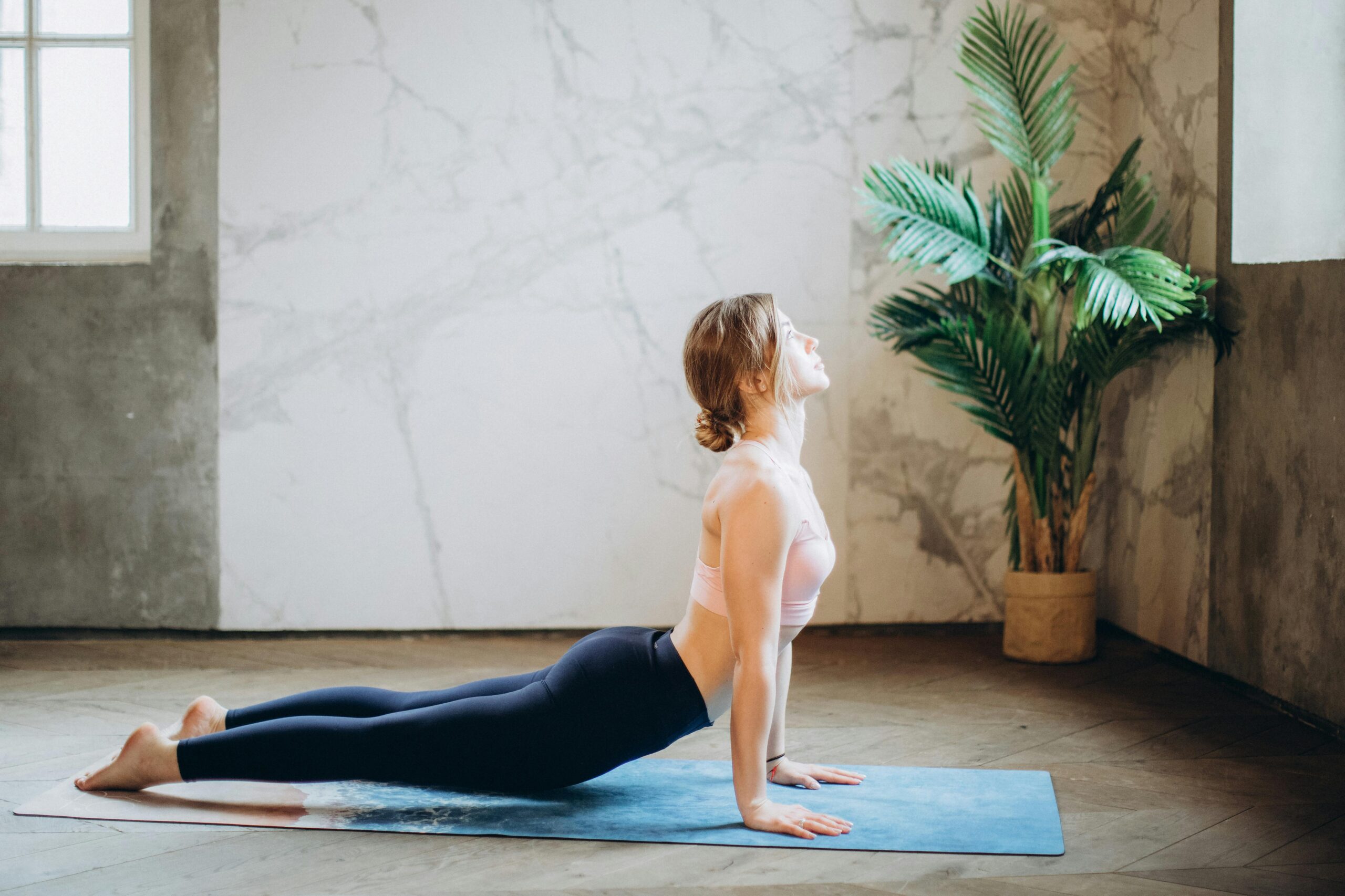 Woman in yoga pose on mat, exercising indoors with natural light and plant decor.